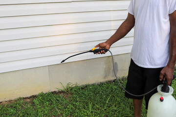 A black man spraying bug spray on the exterior of a house