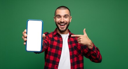 Happy man with beard smiling and pointing at a blank smartphone screen, wearing a plaid shirt on a green background.