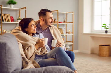 Loving affectionate couple sitting in modern living room wrapped with warm blanket, happy romantic...