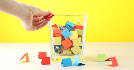 Woman picking red paper piece from glass jar at wooden table against yellow background, closeup