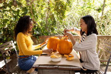 Halloween time, two multiracial female friends carving pumpkins on deck