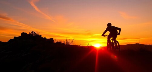 Obraz premium Silhouetted mountain biker descends rugged trail at sunset, vibrant orange sky, downhill, silhouette