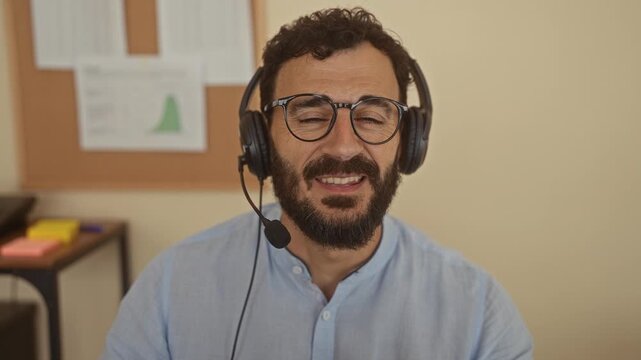 Hispanic man with beard wearing headset smiles and talks in an office setting with charts on the wall and colorful sticky notes in the background.