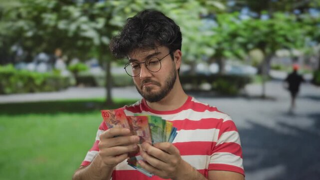 Young man with beard holds swiss francs in a park surrounded by green nature wearing glasses and striped shirt outdoors during daytime