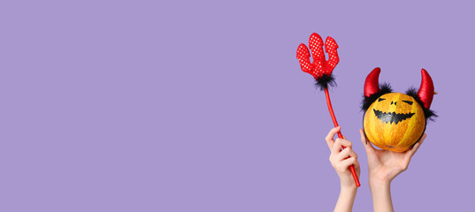 Female hands with pumpkin in red devil horns headband and pitchfork on lilac background. Halloween celebration