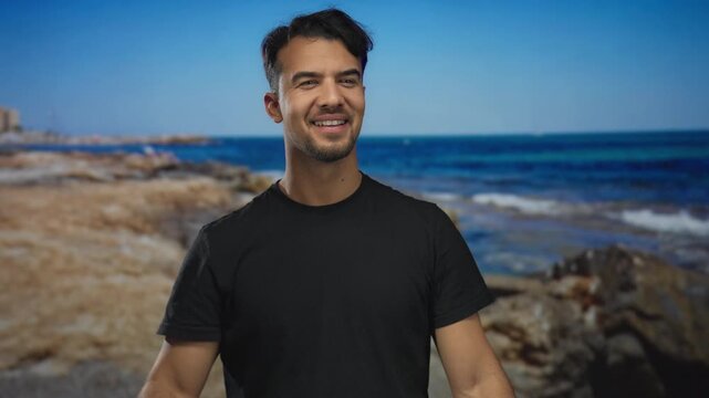 Young hispanic man smiling at a seaside beach with the ocean in the background under a clear blue sky