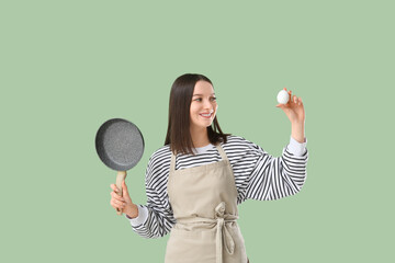 Pretty young woman in apron with chicken egg and frying pan on green background