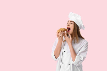 Young female chef eating croissant on pink background