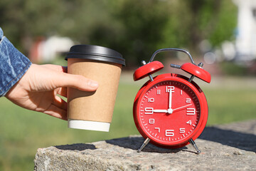 Female hand with coffee cup and red alarm clock in park, closeup