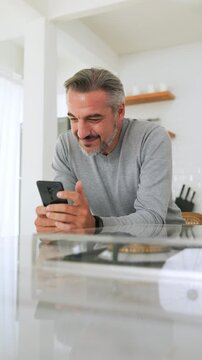 Attractive Middle-aged man using mobile phone or smartphone while standing in kitchen at home.