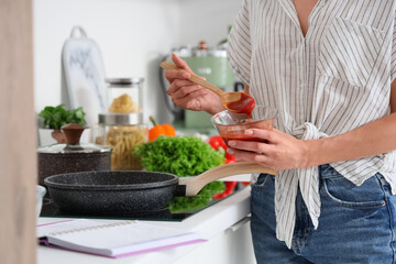 Young woman with tomato sauce and recipe book frying food in kitchen, closeup