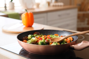 Frying pan with tasty vegetables and spoon on stove in kitchen, closeup