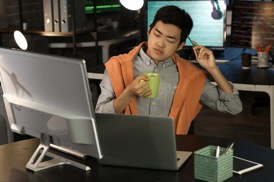 Thoughtful male Asian programmer with coffee in office at night