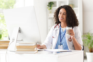 Female African-American medical student with coffee using computer at table in office
