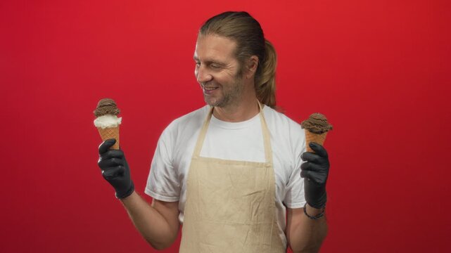 Man wearing apron holding two icecream cones with gloved hands in a bright red studio; happy choice.