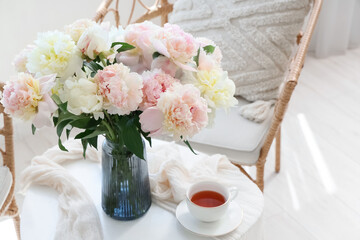 Vase with bouquet of beautiful peonies and cup of tea on table in stylish room, closeup