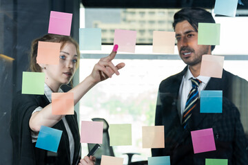 Businesswoman and businessman working together at glass wall with sticky notes during creative team brainstorming in modern office, collaborating on business ideas and strategic direction