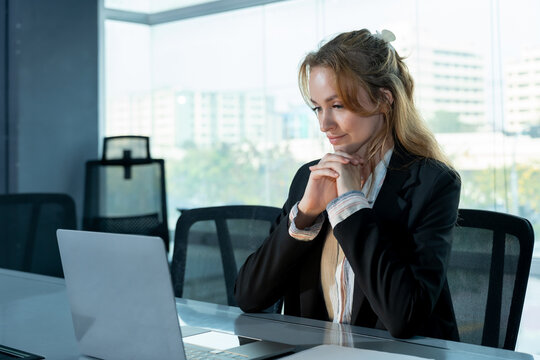Caucasian female executive thinking while looking at laptop screen during solo work session in modern office, planning strategy and analyzing business information for decision making