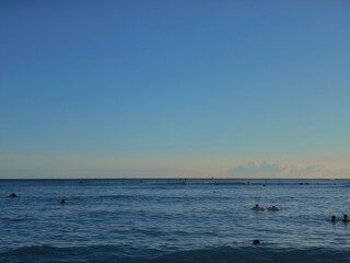 Fototapeta premium The ocean extends to the horizon under a clear, blue sky with a faint golden glow from the setting sun. Dozens of swimmers and surfers float on the gentle waves.