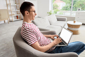 Young man with eyeglasses using blank laptop in armchair at home