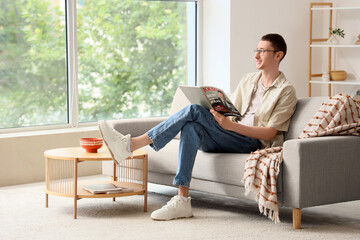 Obraz premium Young man in eyeglasses reading magazine on sofa at home