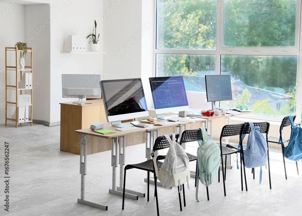 Poster desks with computer monitors and backpacks on chairs in school computer lab