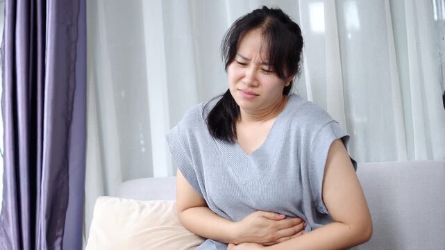 Asian woman experiencing stomach pain while sitting on sofa at home ,signs of indigestion, food poisoning, or menstrual cramps. Holding abdomen due to internal discomfort