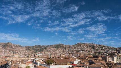 Panorama of the Cuzco city. View from the height. There are many closely built buildings with red tiled roofs. The towers of the Catholic Cathedral are visible. Mountains against a blue sky, clouds. 
