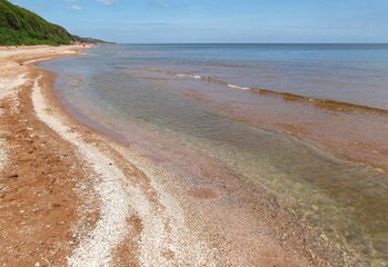Clear shallow water of a sandy sea beach with shell gravel. Green grassy hills and blue sky.