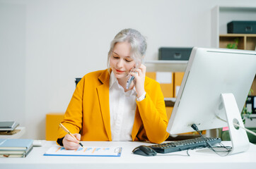 Young female entrepreneur in yellow blazer frustrated during phone call at computer desk in SME office. Customer support issue, complaint, or business stress. Real people.