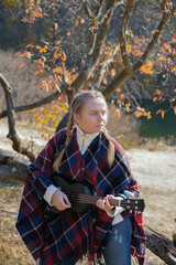 Blonde woman playing guitar ukulele outdoors in autumn forest. Concept of sound therapy, mental...