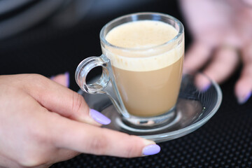 A Close Up Shot of a Delicious Creamy Espresso Macchiato Served in a Clear Glass Cup with a Saucer Held by a Person With Purple Nails