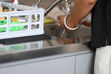Person rinsing dirty dishes in a kitchen sink with running water next to a dish rack with clean dishes and sponges for household chores and cleaning