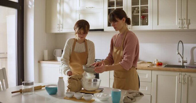 Girl in a beige apron helps her mother sift flour and bakes in a cozy aesthetic kitchen. Team family baking activity in the cozy kitchen at home