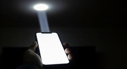 A hand holding a smartphone with a bright screen, illuminated by a ceiling light beam.