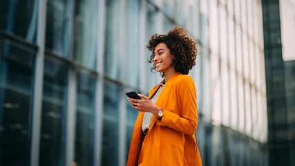Stylish professional woman walking past office buildings, smiling while messaging on phone, urban workday vibe