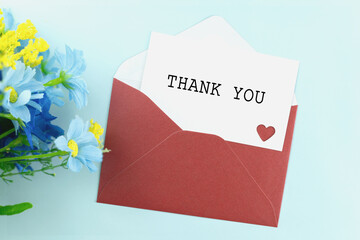  A thank you card is in a red envelope next to a bouquet of blue and yellow flowers on a light blue...