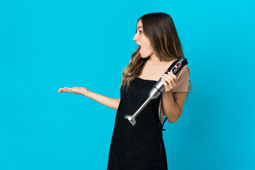 Romanian woman using hand blender isolated on blue background with surprise expression while...