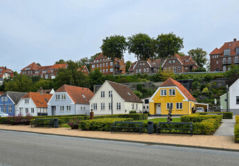 row of charming, colorful houses&mdash;white, yellow, and blue&mdash;lines a quiet residential street, with red-roofed buildings and lush greenery on a hillside in the background, sonderborg, denmark