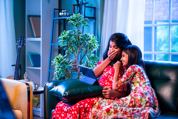 Indian little girls laugh together watching funny video on tablet while relaxing on the sofa