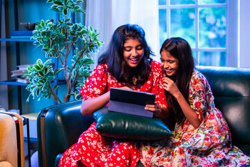 Indian little girls laugh together watching funny video on tablet while relaxing on the sofa