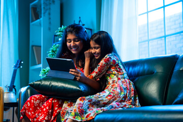 Indian little girls laugh together watching funny video on tablet while relaxing on the sofa