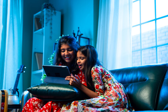 Indian little girls laugh together watching funny video on tablet while relaxing on the sofa - Powered by Adobe