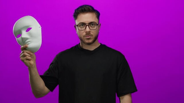 Young man with beard removing white mask against pink wall background in an expressive gesture, showcasing anonymity and identity, isolated in vivid color scheme.