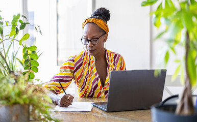 Portrait of confident businesswoman writing notes while using laptop in office
