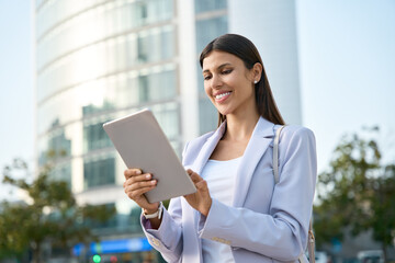 Hispanic smiling woman in suit working using touchpad computer. Successful mature Indian or Latin entrepreneur businesswoman holding digital pc tablet outdoor at business office skyscraper. Copy space
