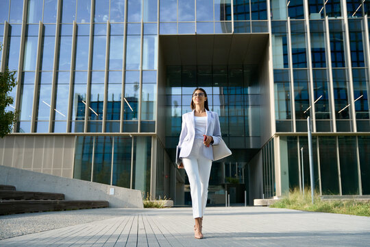 Full body portrait of indian or Latin good looking middle aged leader female businesswoman. Young hispanic woman in stylish business clothes walking outdoor at modern office building. Copy space
