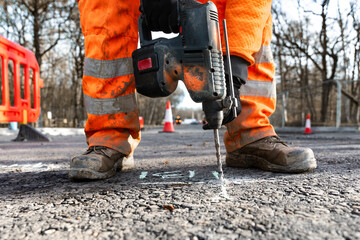 Construction worker in orange clothes and safety boots using cordless drill to break asphalt on road project in rural area