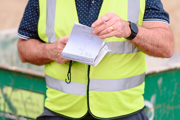 A construction worker in a reflective vest holds a notepad, jotting down details about the ongoing project at a busy construction site