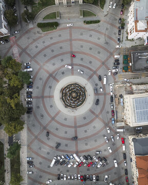 Aerial view of a vibrant circle square bustling with cars around Colchis Fountain, framed by buildings and green trees, Kutaisi, Imereti, Georgia.